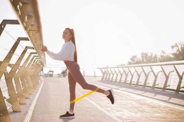Adorable fit woman with long hair dressed in sport apparel doing exercises with rubber band at the bridge in the morning. Space for text - Valokuva, kuva