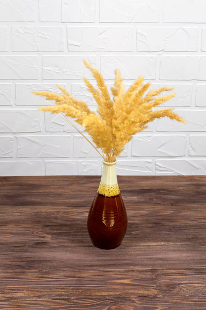 A bouquet of dry spikelets in a brown ceramic vase on a wooden table against a white brick background. Stylish home decor. Modern interior design - Photo, Image