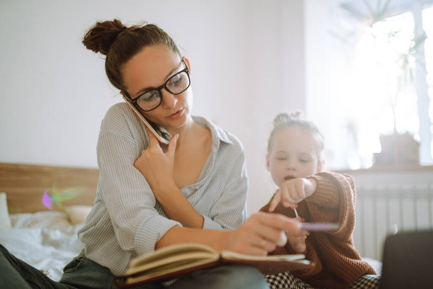 Young beautiful businesswoman talking on mobile phone and working on a laptop. Busy young woman with daughter in home office. Stay at home. Covid-19. - Foto, Imagen