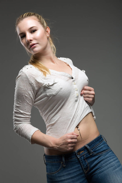 молодая блондинка позирует для фото в серой фотостудии и показывает пупок - Фото, изображение