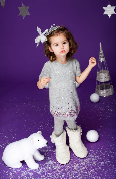 Little cute girl in white winter wreath and with toy polar bear having fun in studio on purple background with silver Christmas trees, stars, snow. Concept of Christmas, New Year's holidays - Photo, Image