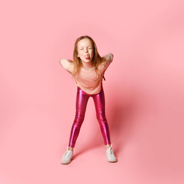 Joyful preschool girl in stylish blouse and leggings is teasing someone leaning forward, grimacing and lolling. Full length shot isolated on pink - Foto, Imagem
