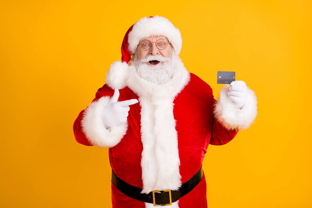 Photo excited grey white beard hair santa claus shopper point finger credit card recommend buy x-mas eve tradition christmas discount wear cap headwear isolated bright shine color background - 写真・画像
