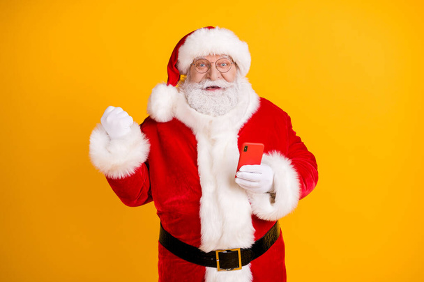 Photo of excited grey white beard hair santa claus use smartphone raise fists celebrate x-mas christmas jolly holly lottery victory wear cap headwear isolated bright shine color background - Photo, Image