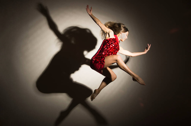 A girl in a red dress performs gymnastic jumps, a shadow from a figure appears on a white background. Studio photography. - Фото, зображення
