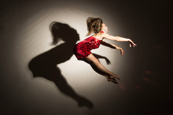 A girl in a red dress performs gymnastic jumps, a shadow from a figure appears on a white background. Studio photography. - Foto, Bild