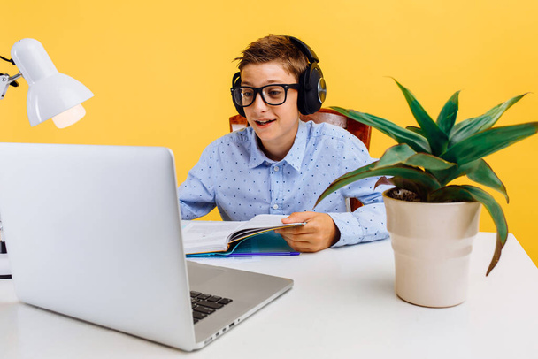 Online lessons for children. Home schooling and distance education for children. schoolboy sitting at a table reading a book, learning by video link with a teacher, on a yellow background - Photo, Image
