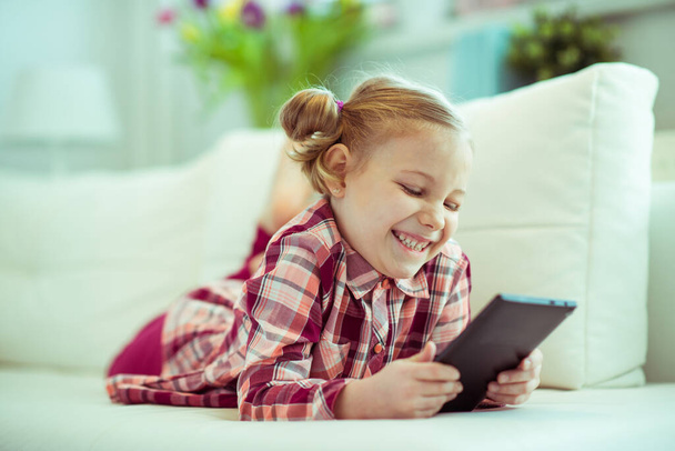 Portrait of cute little child girl laying on sofa with tablet at digital homeschooling - Фото, изображение