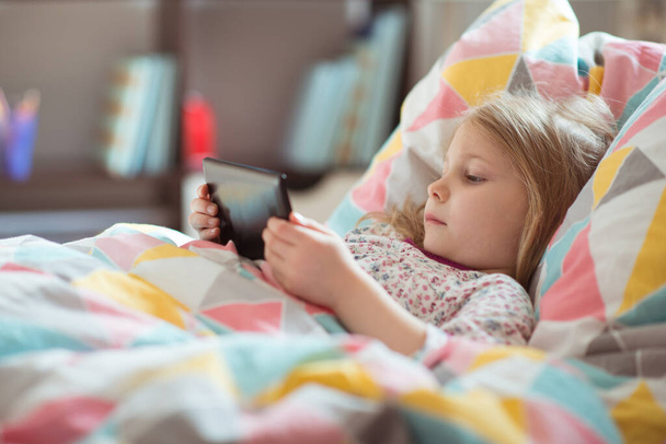 Portrait of cute little blonde preschool child girl laying ill in bed with tablet at homeschooling - Foto, afbeelding
