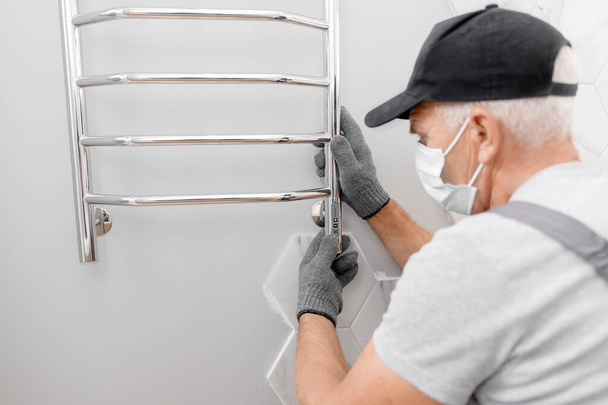 Plumber in medical mask installs electric heated towel rail in bathroom - Photo, Image