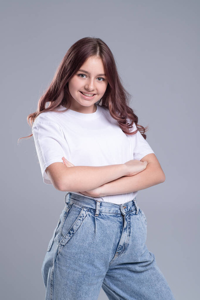 Portrait of nice caucasian teenage girl in white t-shirt and jeans posing on gray background with crossed arms. Self confidence concept - Photo, Image