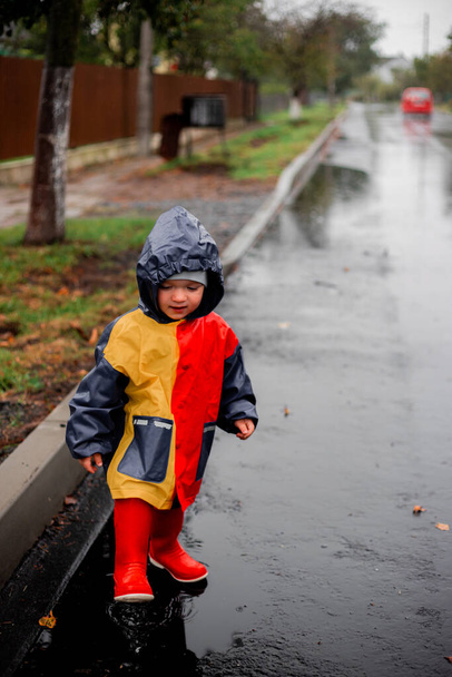 Małe dziecko, samotne w deszczu, biegnie na kalyuzh.W płaszczu przeciwdeszczowym w kolorze niebieskim, czerwonym i pomarańczowym oraz czerwonych butach. Szczęśliwe dziecko. Beztroskie dzieciństwo. Zła pogoda. Lubię deszcz. mgła - Zdjęcie, obraz