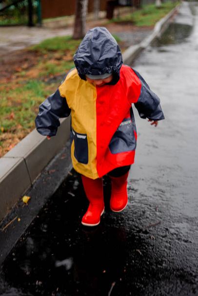 Yağmurda tek başına koşan küçük bir çocuk kalyuzhla koşar. Mavi, kırmızı ve turuncu renkli ve kırmızı çizmeli bir yağmurlukla. Mutlu çocuk. Tasasız çocukluk. Kötü hava. Yağmuru severim. sis. - Fotoğraf, Görsel