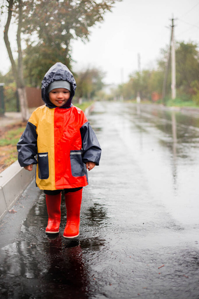 Uma criança pequena, sozinha na chuva, corre em kalyuzh.Em uma capa de chuva nas cores azul, vermelho e laranja e botas vermelhas. Criança feliz. Infância despreocupada. Mau tempo. Eu gosto de chuva. nevoeiro - Foto, Imagem