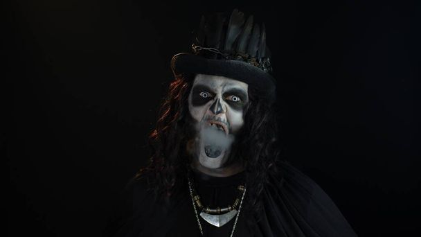 Μοχθηρός άνθρωπος με αποκριάτικο σκελετό μακιγιάζ εκπνέοντας τσιγάρο από το στόμα του και χαμογελώντας - Φωτογραφία, εικόνα