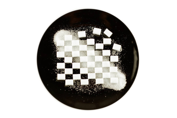 cubetti di zucchero raffinato deposto in un modello su un piatto nero, zucchero di cristallo sparso sulla parte superiore, isolato su bianco, vista frontale, blocchi di modello parzialmente distrutto - Foto, immagini