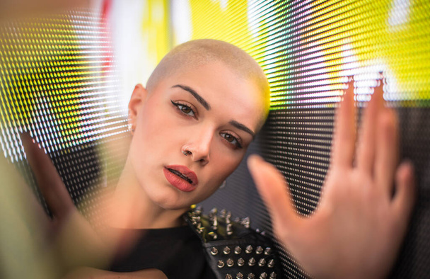 Image of a beautiful young woman posing against a led panel. Shaved head teenager with alternative look making urban portraits - Zdjęcie, obraz