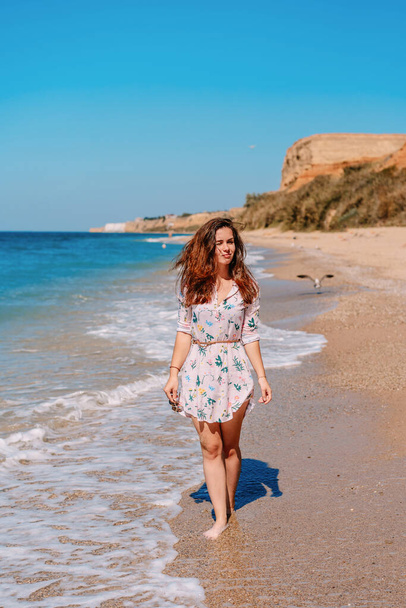 A young woman in a dress and long hair walks along the coast by the sea with waves, the concept of a relaxing holiday - Photo, image