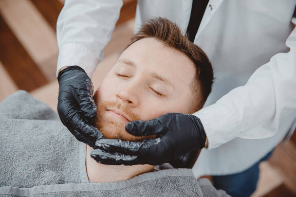 Barber kapper brengt schuim en baard scheerlotions aan op mannelijke klant. Crème en olie huidverzorging concept - Foto, afbeelding