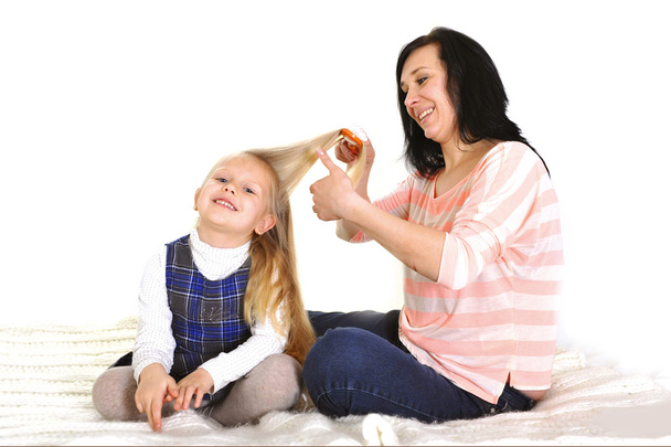 Young beautiful mother with hairbrush brushing daughter blonde hair - Photo, image