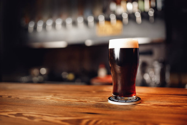 Verre d'artisanat de bière sombre avec mousse sur comptoir de bar en bois, fond flou pub - Photo, image