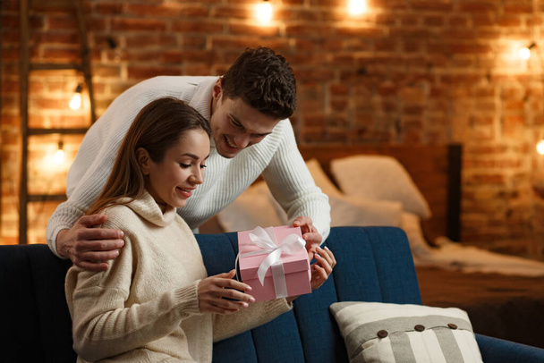 Handsome man presenting a gift to his beautiful girl and smiling. Beautiful young couple at home enjoying spending time together. Winter holidays, Christmas celebrations, New Year concept - Φωτογραφία, εικόνα