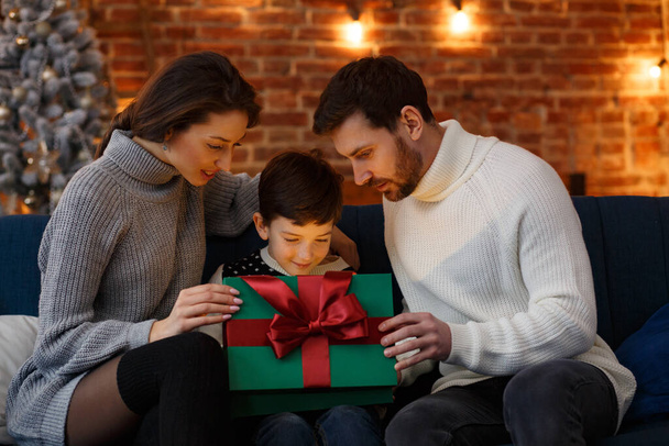 Vanhemmat esittelevät lahjapakkauksen söpölle pojalle. Joululapsia, lapsi, muotokuva. Onnellinen perhe viettää jouluaamun yhdessä. Talvilomat, Joulujuhlat, Uudenvuoden konsepti - Valokuva, kuva