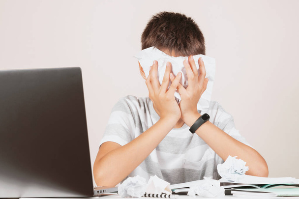 Το θλιμμένο παιδί κάθεται σε ένα τραπέζι με υπολογιστή στο σπίτι. Έχει προβλήματα με την εργασία. Το παιδί απογοητεύτηκε και έκρυψε το πρόσωπό του με ένα τσαλακωμένο χαρτί. - Φωτογραφία, εικόνα