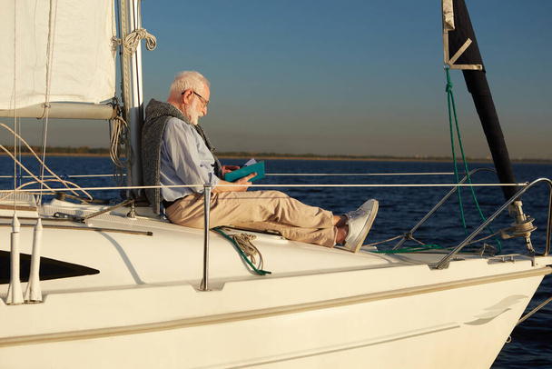 Hombre mayor relajado lectura libro mientras está sentado en el lado de la cubierta de velero o yate flotando en el mar azul calma en un día soleado - Foto, imagen