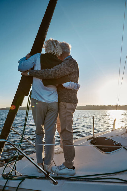 Vue arrière du couple de personnes âgées embrassant et regardant l'horizon, profitant d'un coucher de soleil incroyable tout en se tenant sur le côté du voilier ou du pont de yacht flottant en mer - Photo, image