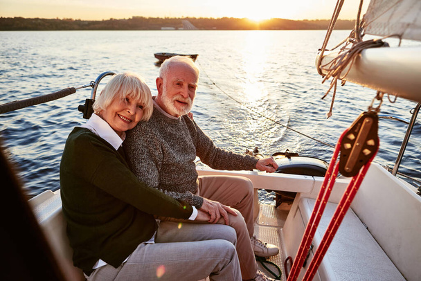 Heureux couple de personnes âgées amoureux, homme et femme âgés tenant la main, embrasser et passer du temps ensemble tout en étant assis sur le côté du pont de yacht flottant dans la mer par une journée ensoleillée - Photo, image