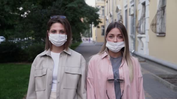 Portret dwóch pięknych młodych kobiet noszących ochronne maski medyczne i stojących na ulicy. Bezpieczne i szczęśliwe kobiety ćwiczące dystans społeczny, kwarantannę. Zamazane tło miasta - Materiał filmowy, wideo
