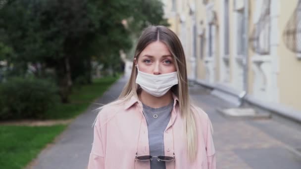 Close-up van het gezicht van blanke jonge mooie vrouw met respiratoire virus dragen masker op straat buiten in de stad. Portret van een beschermd triest meisje dat naar de camera kijkt. Gezondheids- en veiligheidsconcept. - Video