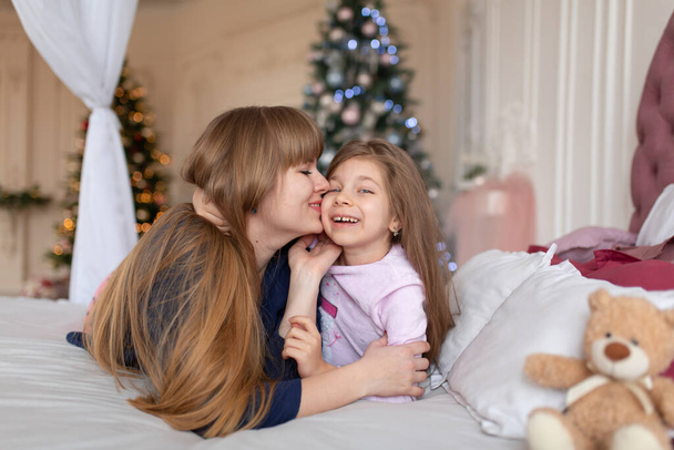 ベッドで寝そべっている間、女の子はお母さんと遊ぶ時間を過ごす。クリスマスの話。幸せな子供時代. - 写真・画像