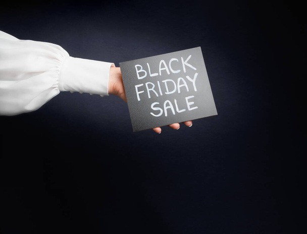 ένα γυναικείο χέρι σε ένα λευκό πουκάμισο κρατά μια πινακίδα με τις λέξεις "μαύρη Παρασκευή πώληση", σε μαύρο φόντο - Φωτογραφία, εικόνα
