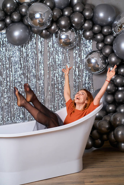 The beautiful red-haired girl rejoices sitting dressed in the bath. against the background of tinsel and balloons. Success concept. - Foto, imagen