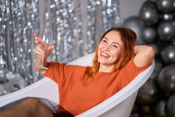 The beautiful red-haired girl rejoices sitting dressed in the bath. against the background of tinsel and balloons. Success concept. - 写真・画像