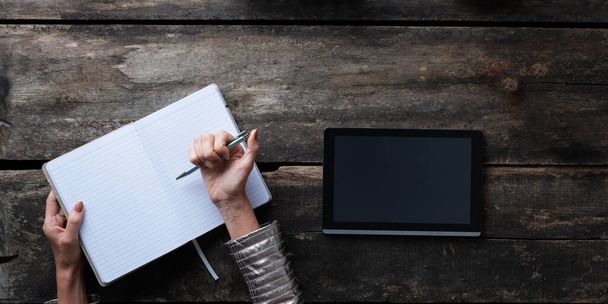 Κάτοψη μιας γυναίκας που ετοιμάζεται να κρατήσει σημειώσεις σε ένα κενό ανοιχτό σημειωματάριο δίπλα σε ένα ψηφιακό tablet. - Φωτογραφία, εικόνα