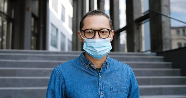 Portrait of young handsome male student in medical mask and glasses standing outdoors and looking at camera. Coronavirus pandemic concept. Caucasian man at street in eyeglasses and protection. - Photo, Image