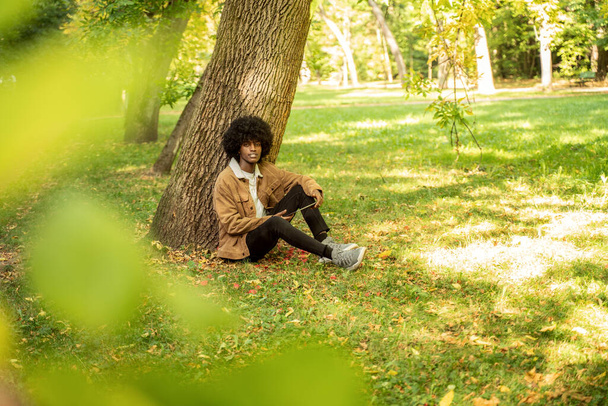 Young African American man using tablet in park. - 写真・画像