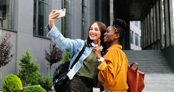 Mixed-races young pretty females smiling cheerfully to camera and taking selfie photo with mobile phone camera at city street. Multi ethnic beautiful happy women students making photos with smartphone - 写真・画像