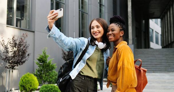 Mixed-races young pretty females smiling cheerfully to camera and taking selfie photo with mobile phone camera at city street. Multi ethnic beautiful happy women students making photos with smartphone - Fotó, kép