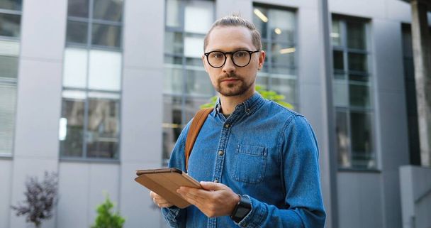 Portrait of Caucasian young man in glasses tapping on tablet device and smiling to camera at city street. Handsome male texting message and browsing on gadget computer outdoors. Gadget user concept. - 写真・画像