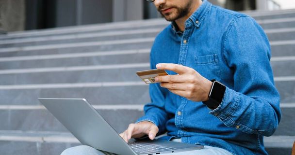Caucasian young man in glasses and blue jeans shirt using credit card and shopping online on laptop. Good-looking male in eyeglasses typing on computer and buying in internet. Buyer at street. - Foto, Imagem