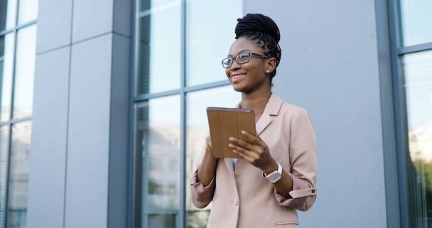 African American young businesswoman in glasses tapping and scrolling on tablet device. Woman texting message on computer outdoors. Confident female using gadget and chatting. - 写真・画像