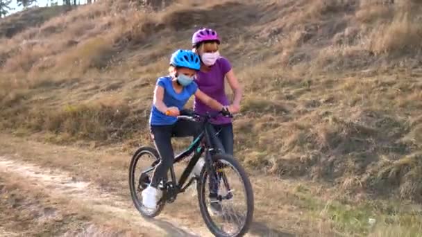 Genç bir anne kızına tıbbi maskeli bisiklet sürmeyi ve tepeye yakın bir yerde bisiklet kaskı takmayı öğretir. Covid-19 koronavirüs enfeksiyonundan korunma.  - Video, Çekim