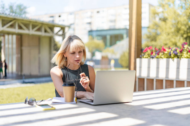 Mulher adulta em óculos com videochamada com laptop fora no parque. Feliz e sorridente sênior trabalhando e bebendo café. Usando o computador. Educação à distância online e lojas online - Foto, Imagem