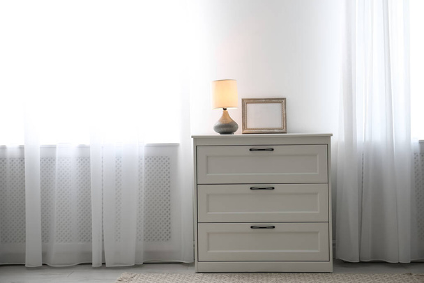 Modern chest of drawers with lamp near window indoors - Foto, Bild