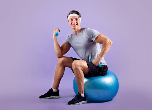 Retrato de comprimento total de cara jovem em sportswear sentado na bola de fitness e levantando halteres no fundo violeta - Foto, Imagem