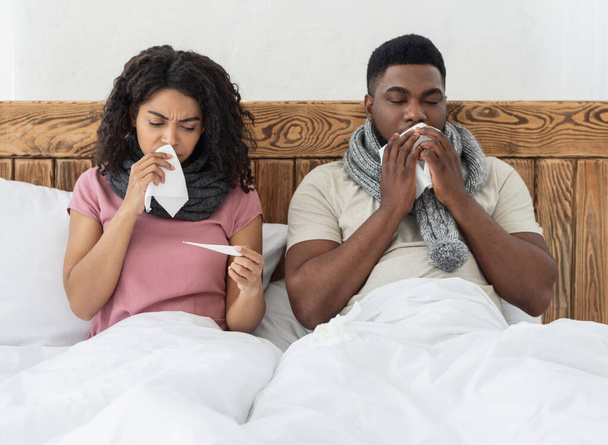 Sick african couple sitting in bed, measuring fever, sneezing noses - Foto, Imagen